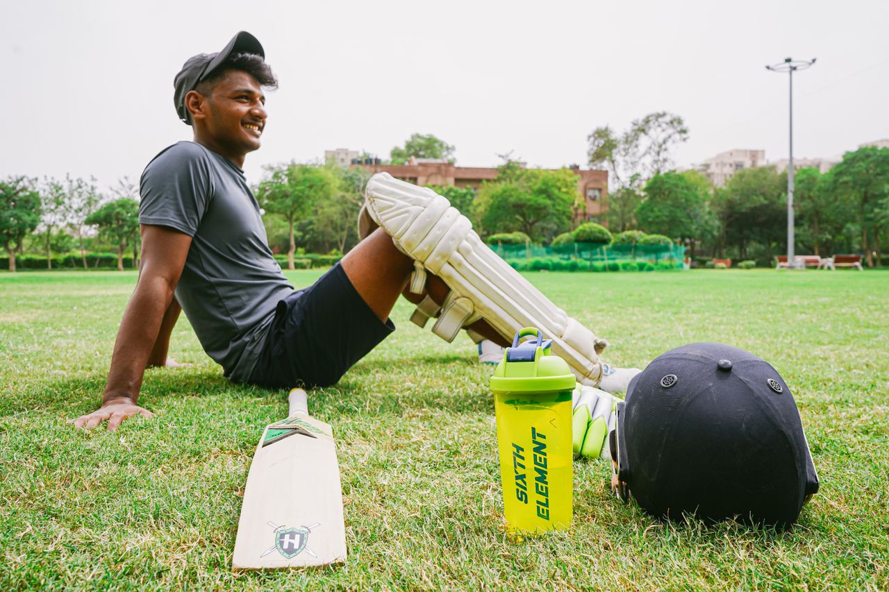 hero-services Side view of full body happy sportsman wearing sportswear and cricket pads resting on lawn beside cricket bat and helmet and sport water bottle